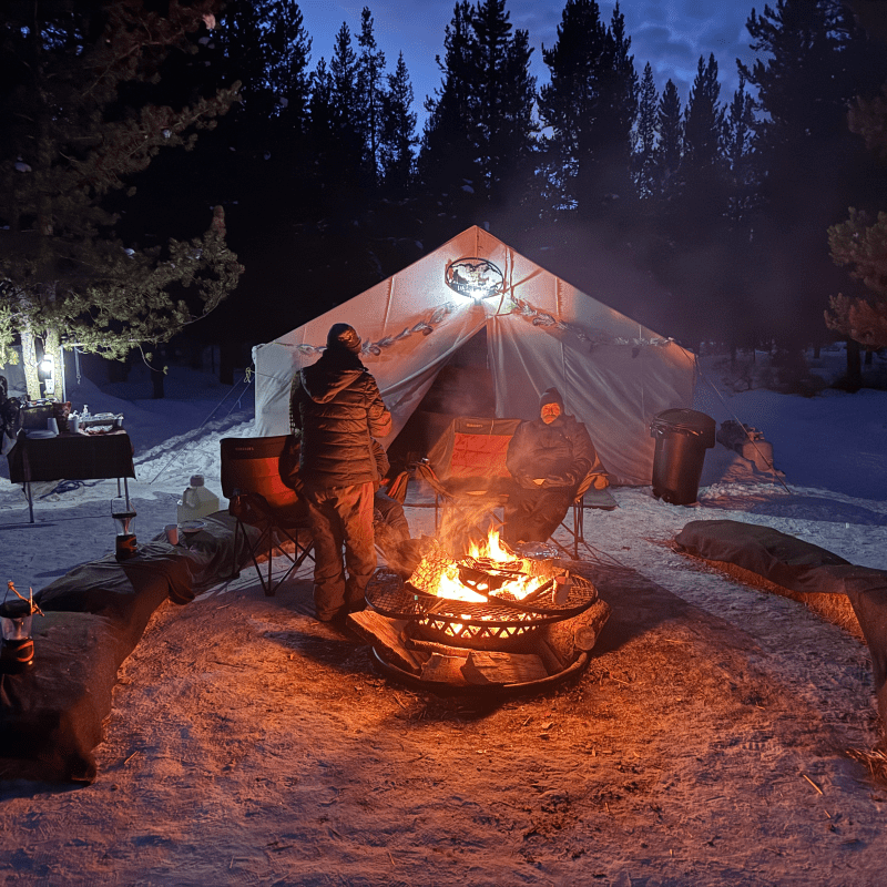 Two people around a campfire in front of a tent at night in a snowy forest.